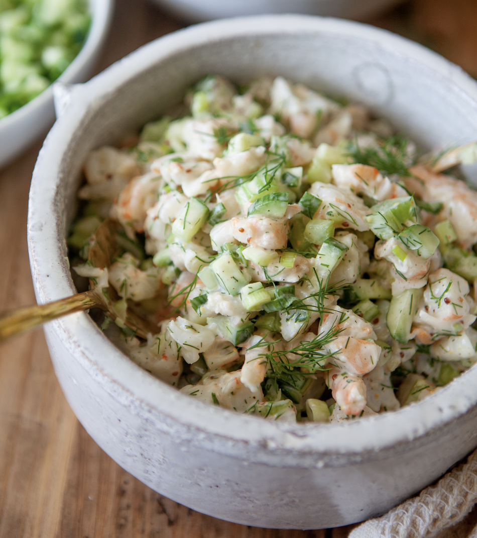 A ceramic bowl filled with Cold Shrimp Salad featuring chopped celery, fresh dill, and a creamy dressing, with a gold spoon inside.