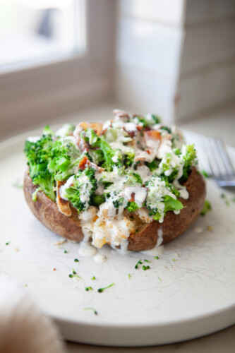 Broccoli cheese stuffed potato topped with steamed broccoli, bacon, and sliced chicken, drizzled with creamy herb sauce on a white plate.