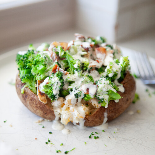 Broccoli cheese stuffed potato topped with steamed broccoli, bacon, and sliced chicken, drizzled with creamy herb sauce on a white plate.