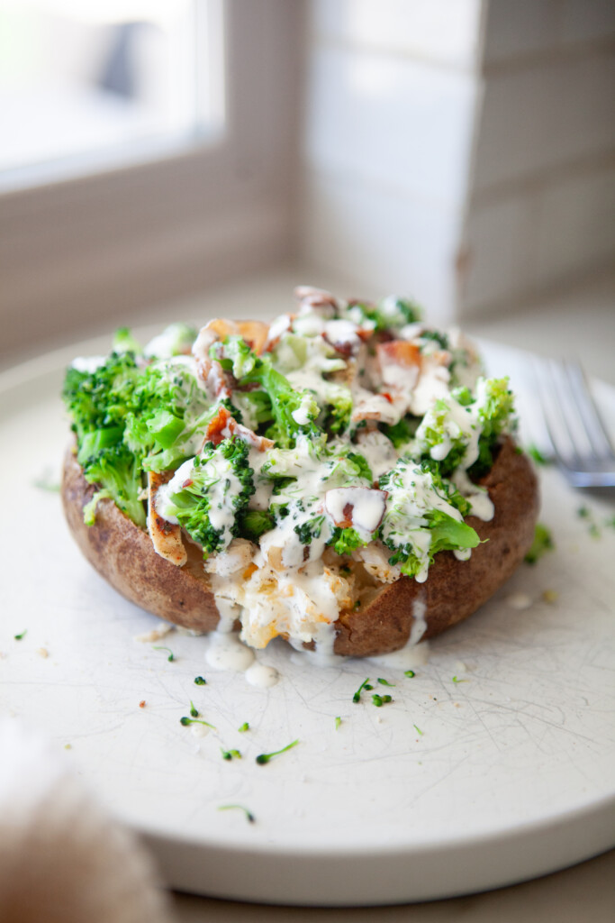 Broccoli cheese stuffed potato topped with steamed broccoli, bacon, and sliced chicken, drizzled with creamy herb sauce on a white plate.