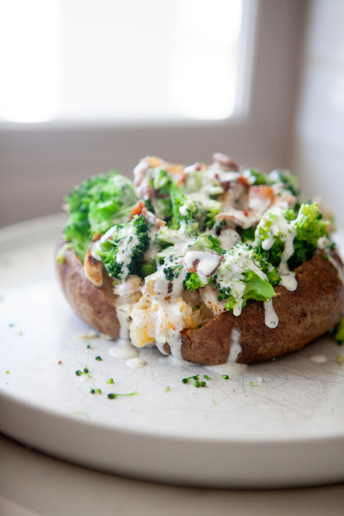 Close-up of a broccoli cheese baked potato with melted cheddar inside, topped with steamed broccoli, bacon, sliced chicken, and creamy herb sauce dripping onto a white plate.