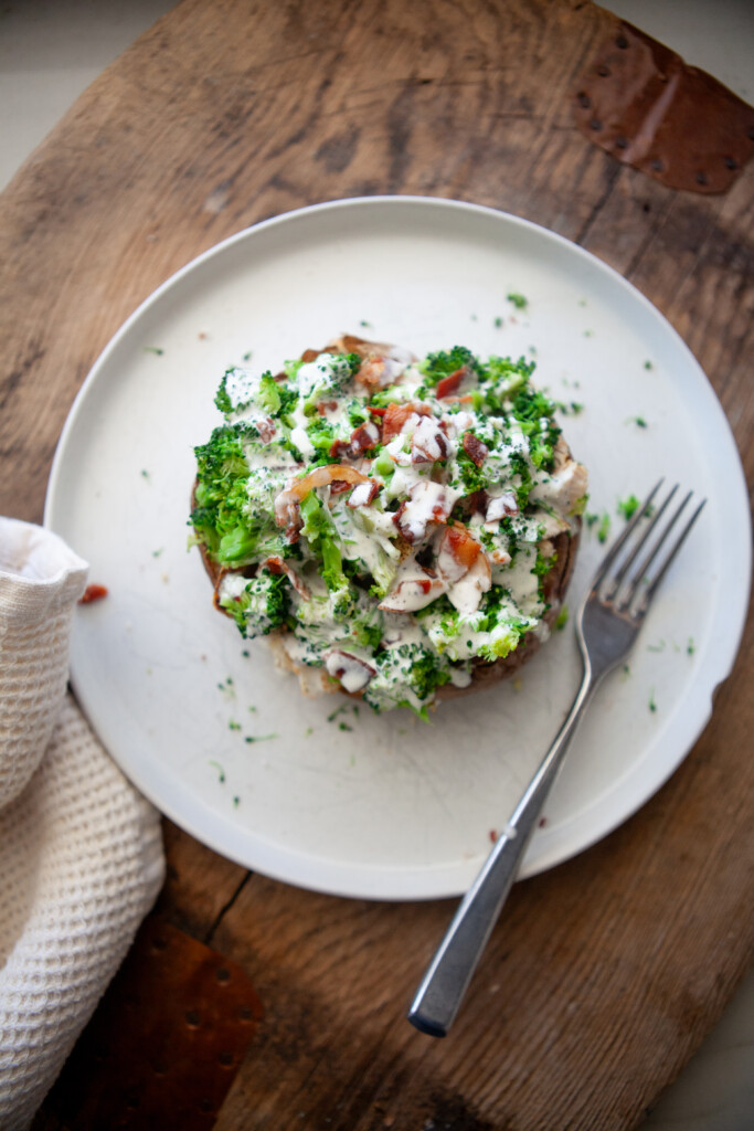 Overhead view of a broccoli cheese baked potato topped with steamed broccoli, crumbled bacon, and creamy sauce on a white plate with a fork.