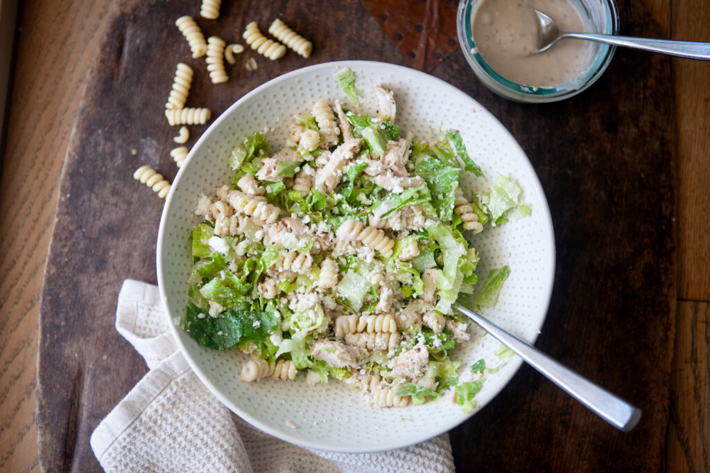 Caesar pasta salad in a white bowl on a wooden table beside a small dish of dressing and scattered dry noodles.