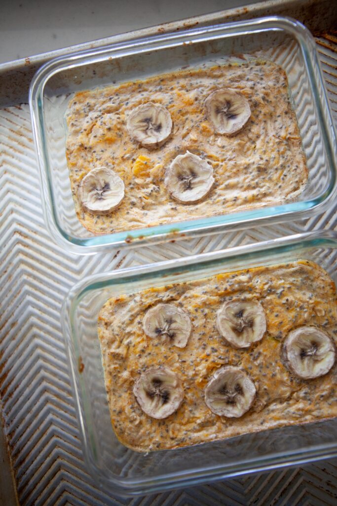 Two banana butternut baked oat bowls in glass containers on a baking sheet, fresh out of the oven with banana slices on top