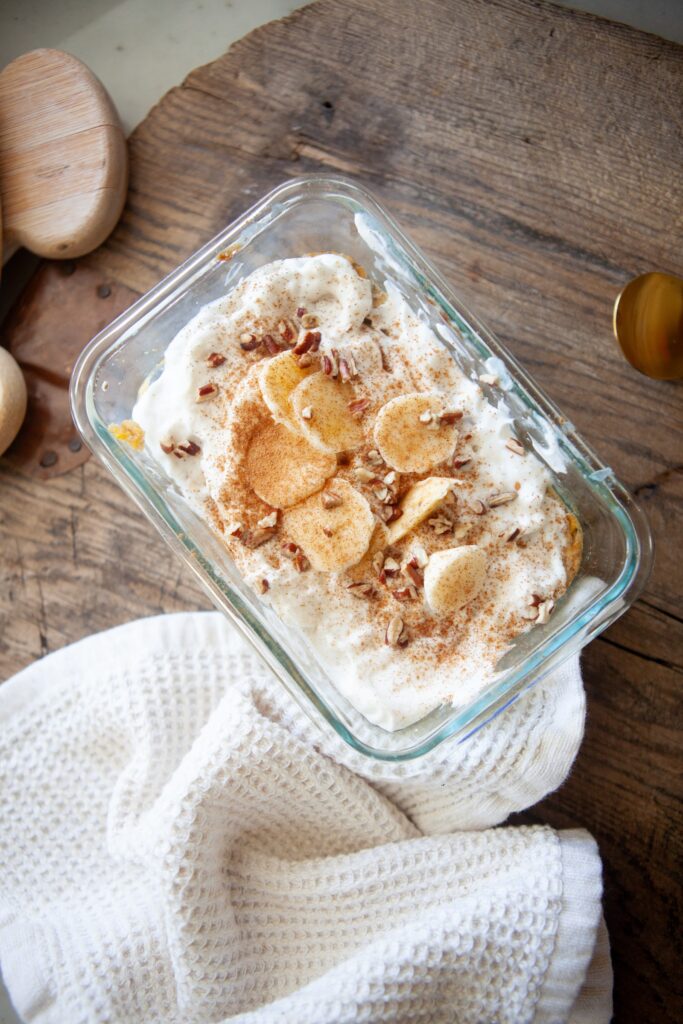 Overhead view of a high protein banana butternut baked oat bowl in a glass container topped with whipped topping, banana slices, cinnamon, and chopped nuts