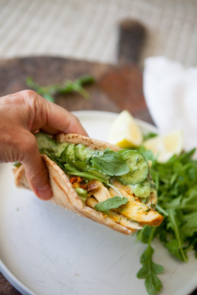 Hand holding a folded chicken shawarma pita filled with creamy cucumber salad and fresh greens over a white plate