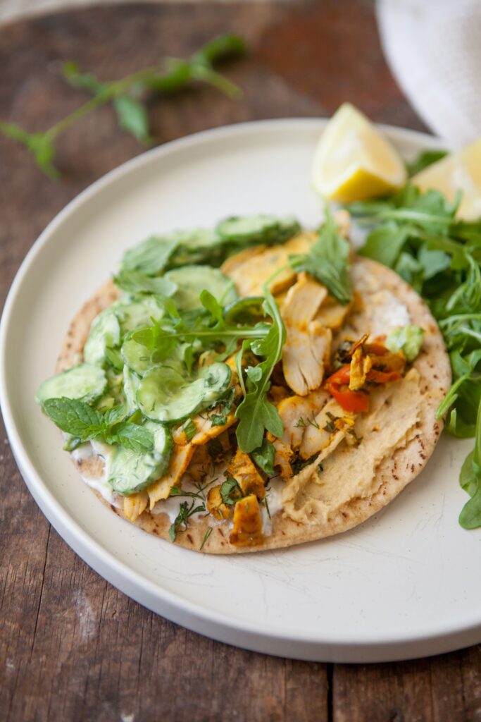 Open-faced chicken shawarma pita spread with hummus and yogurt, topped with creamy cucumber salad and arugula, with lemon wedges on the side