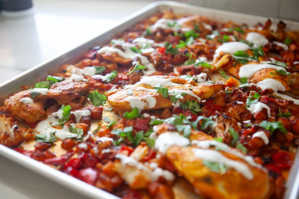 Angled view of an XL sheet pan of Moroccan chicken and vegetables topped with yogurt and cilantro