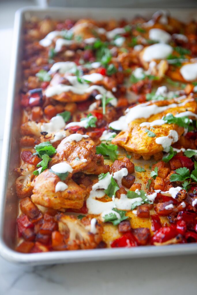 Overhead view of a Moroccan chicken sheet pan dinner with yogurt-marinated chicken thighs, roasted butternut squash, cauliflower, red peppers, yogurt drizzle, and cilantro