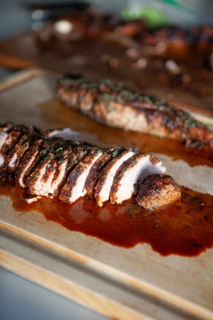 Two oven baked BBQ pork tenderloins on a cutting board, one sliced and one whole, coated in ranch seasoning and parsley