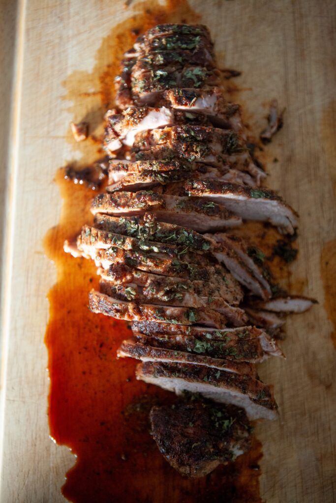 Overhead view of sliced oven baked BBQ pork tenderloin on a wooden cutting board with BBQ sauce juices and parsley
