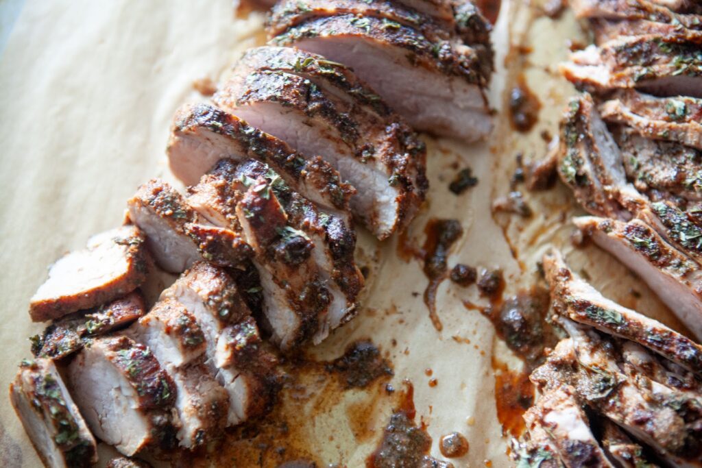 Two sliced BBQ pork tenderloins side by side on parchment paper with seasoning and pan juices