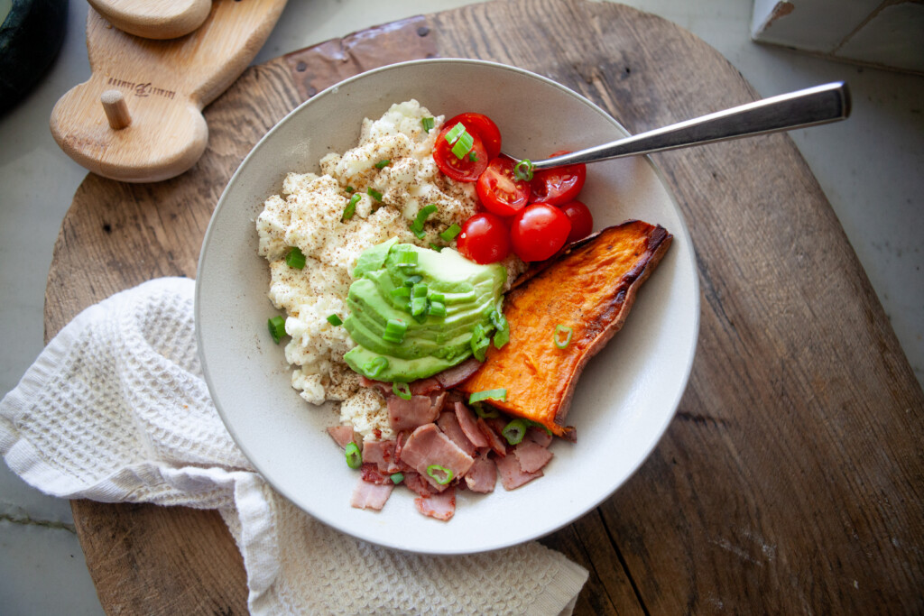 A breakfast bowl containing scrambled egg whites, sliced avocado, cherry tomatoes, a wedge of sweet potato, and chopped ham, topped with green onions and a fork.