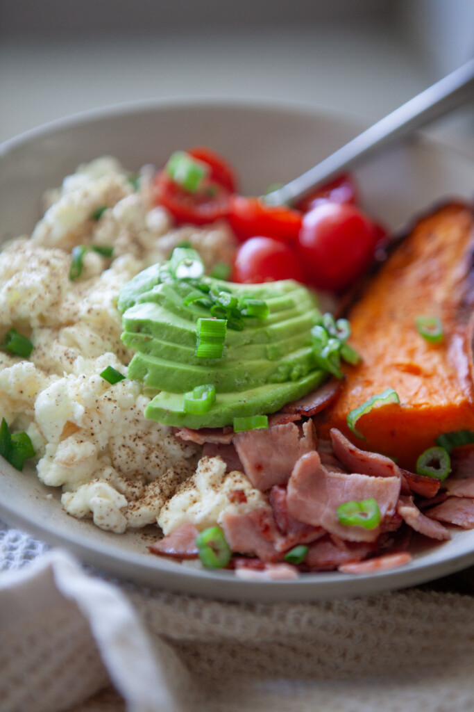 A bowl containing scrambled egg whites, sliced avocado, sliced ham, cherry tomatoes, and a roasted sweet potato wedge.